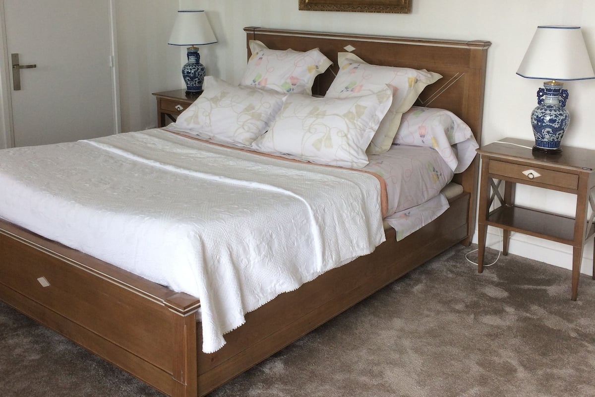 A neatly made bed with a wooden frame, two bedside tables with lamps, a door, and a painting on the wall.