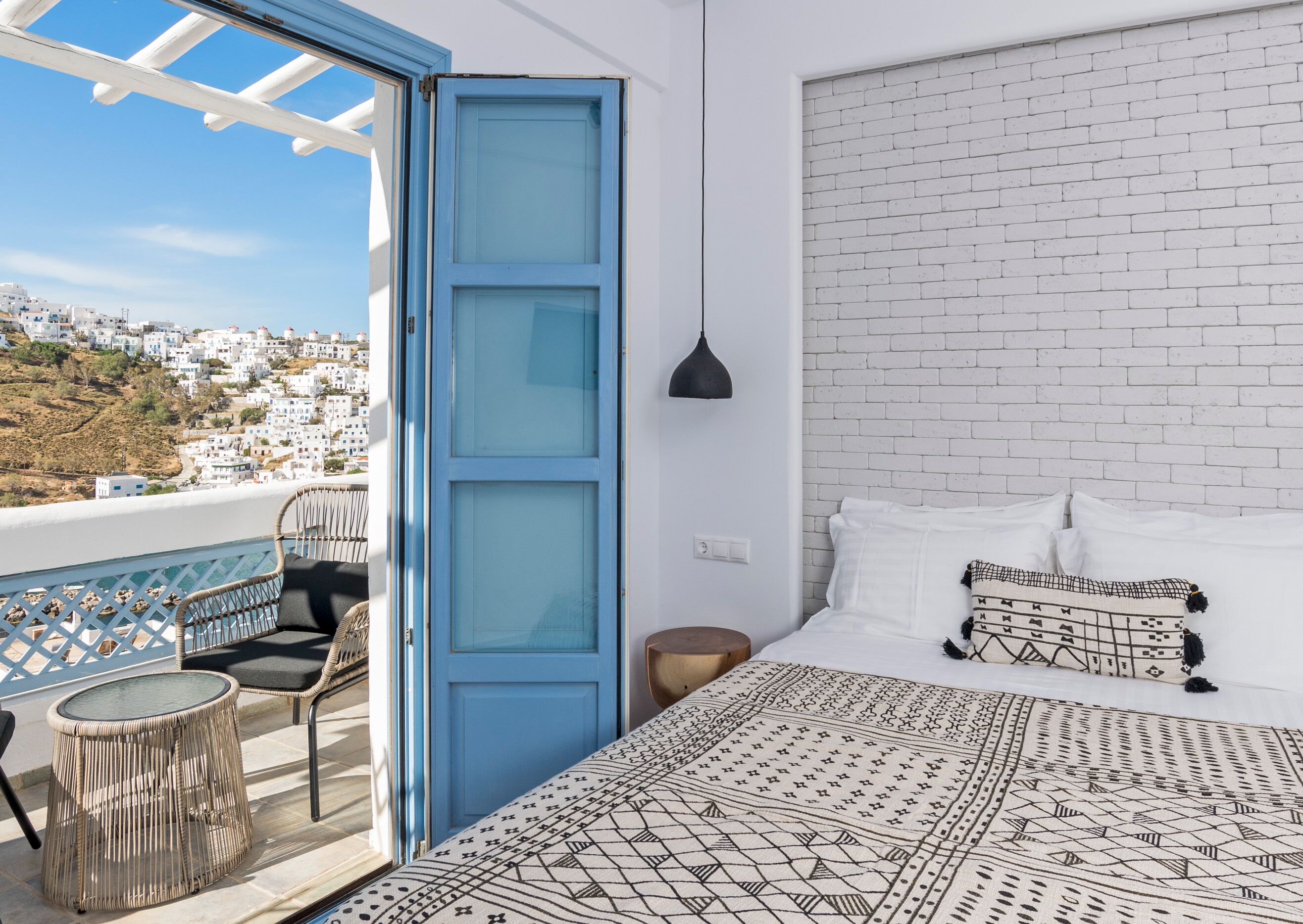 Une chambre avec une tête de lit en briques blanches, une couvre-lit à motifs, une porte bleue, un fauteuil en osier et une petite table ronde.
