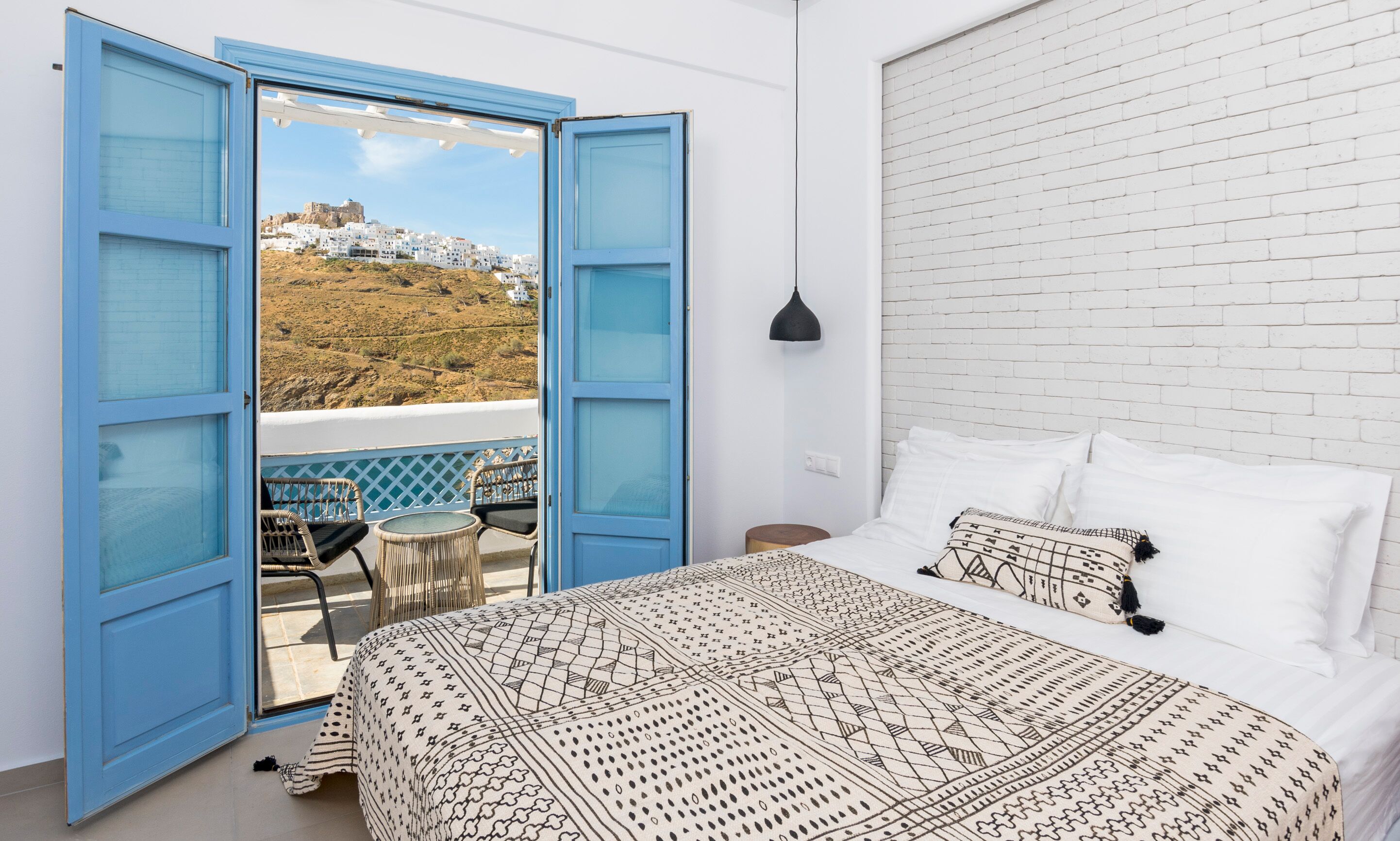 Un dormitorio con cama, almohadas blancas y una colcha estampada. Hay un balcón con puertas azules y vistas a un edificio blanco en una colina.