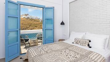 Un dormitorio con cama, almohadas blancas y una colcha estampada. Hay un balcón con puertas azules y vistas a un edificio blanco en una colina.