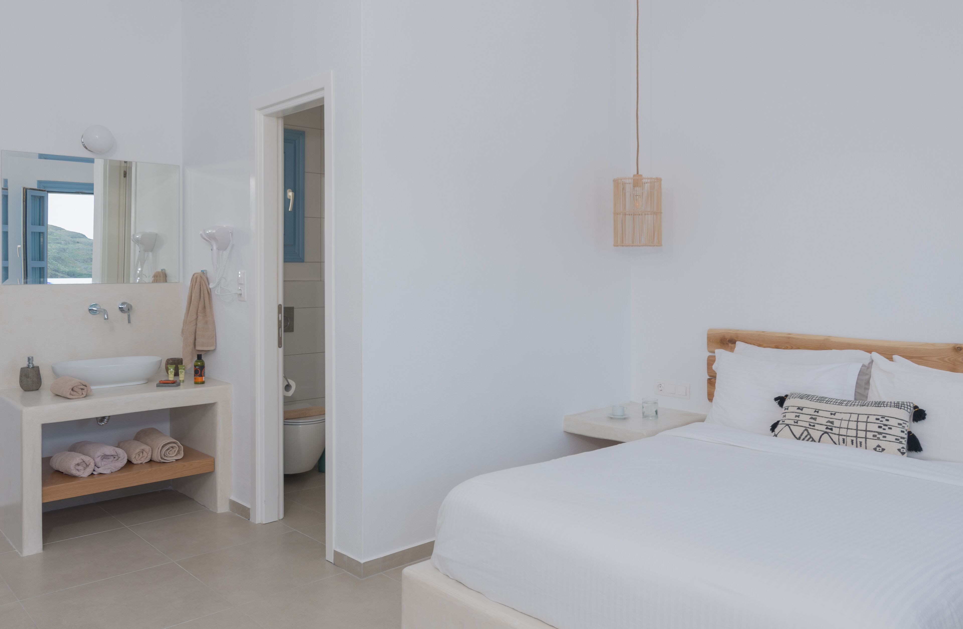 Habitación de hotel minimalista con una cama grande, una mesita de noche, un toallero y un cabecero de madera. Incluye un lavamanos blanco con espejo.