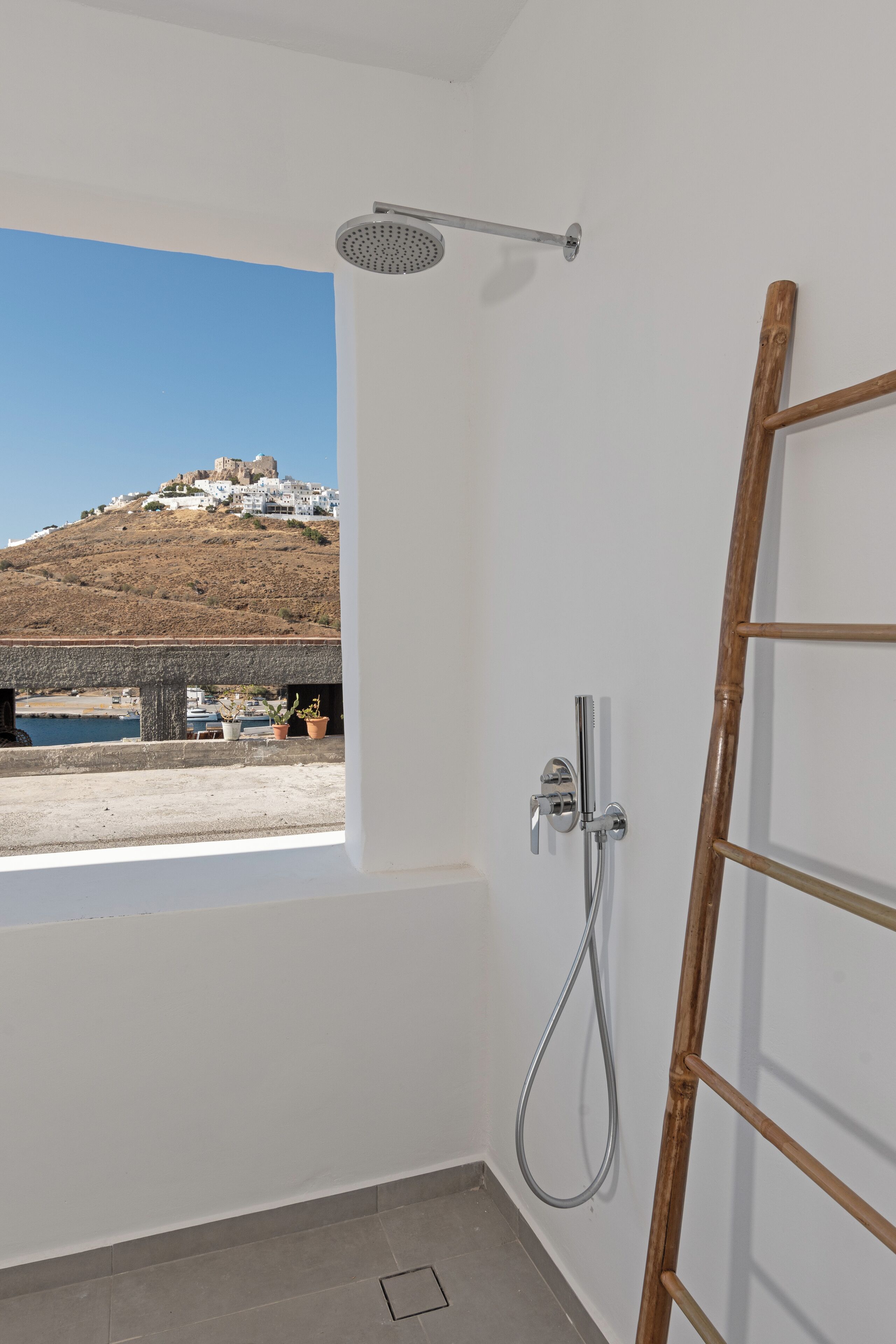 A bathroom with a shower, a window with a view of a hill and buildings, and a wooden door.
