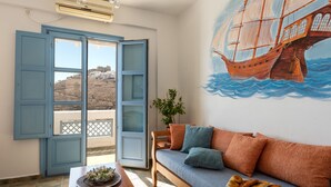 A room with a wooden sofa, a table with a plant and a plate of pastries, a mural of a sailing ship, and a view through a door to a balcony with a white building. - Ihthioessa Boutique Hotel (Astypalaia)