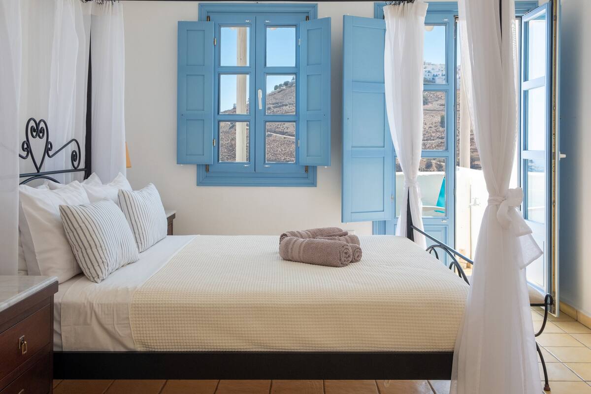 Une chambre à coucher avec un lit, des rideaux blancs, une vue sur l’extérieur et une fenêtre avec des volets bleus.