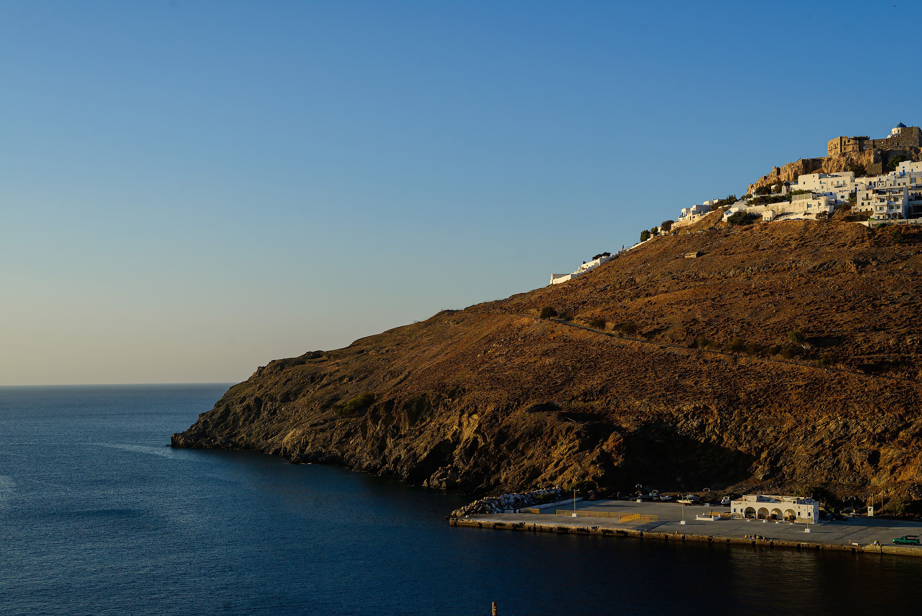 A coastal town with white buildings on a hillside, a rocky cliff, and a calm sea.