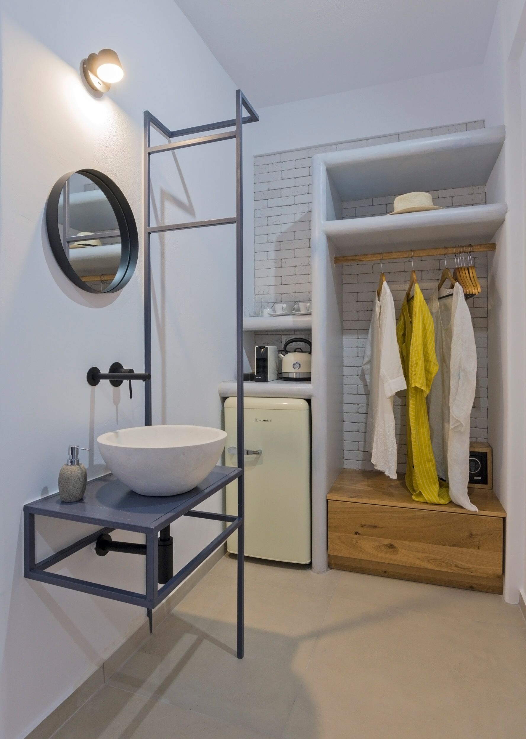 Een moderne badkamer met een witte wastafel, een ronde spiegel, een vrijstaand plankje, een kleine witte koelkast en een houten wastafelmeubel met daaraan hangende kleding.