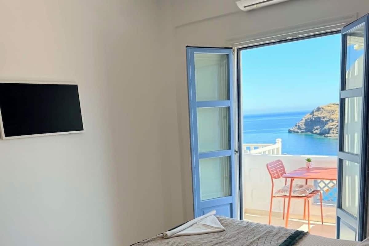 Une chambre avec un lit, une télévision, un balcon avec une table et des chaises, et une vue sur la mer.