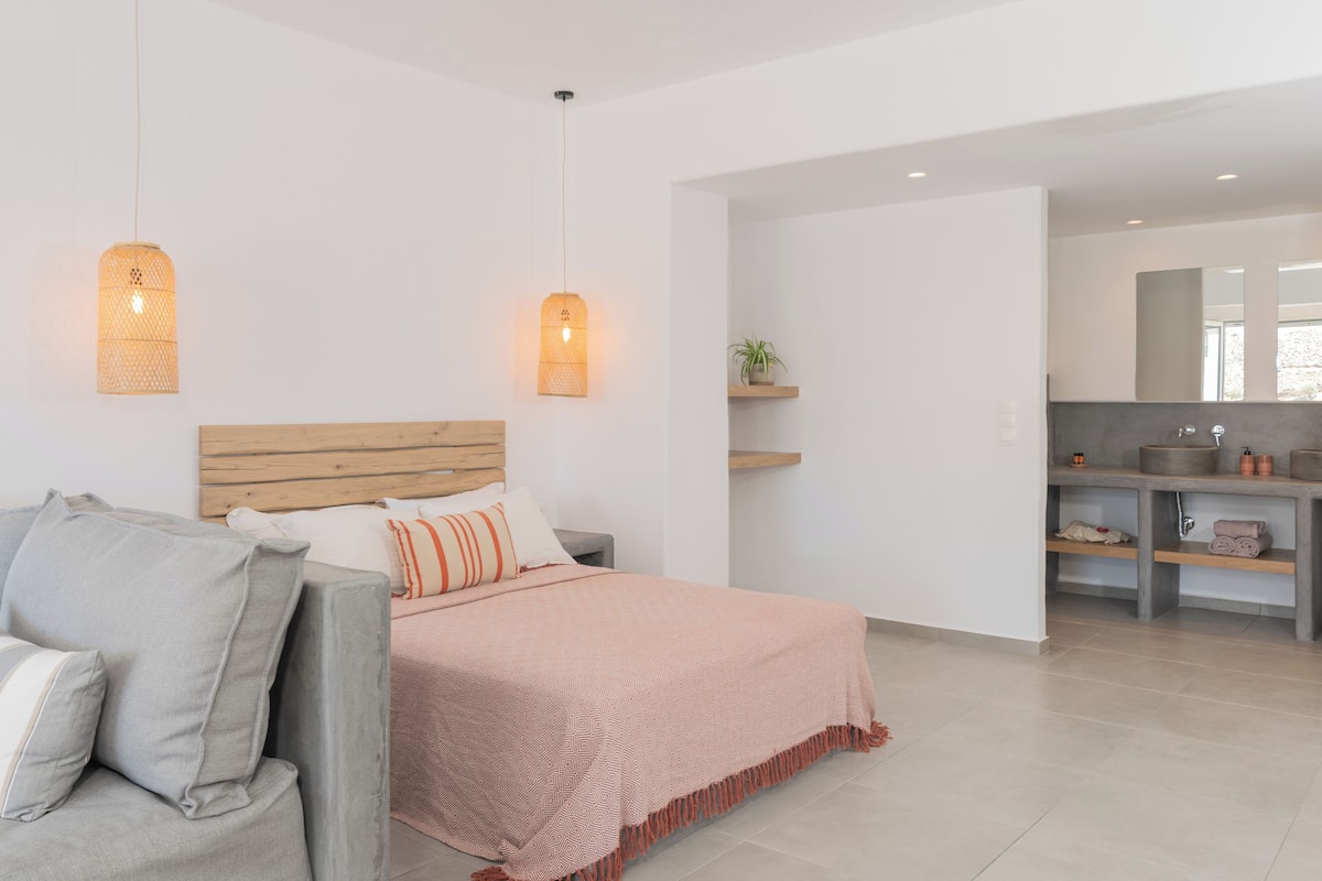 Une chambre moderne avec une tête de lit en bois, un canapé gris et une couvre-lit rose. On aperçoit une petite plante sur une étagère et, au fond, une salle de bain.