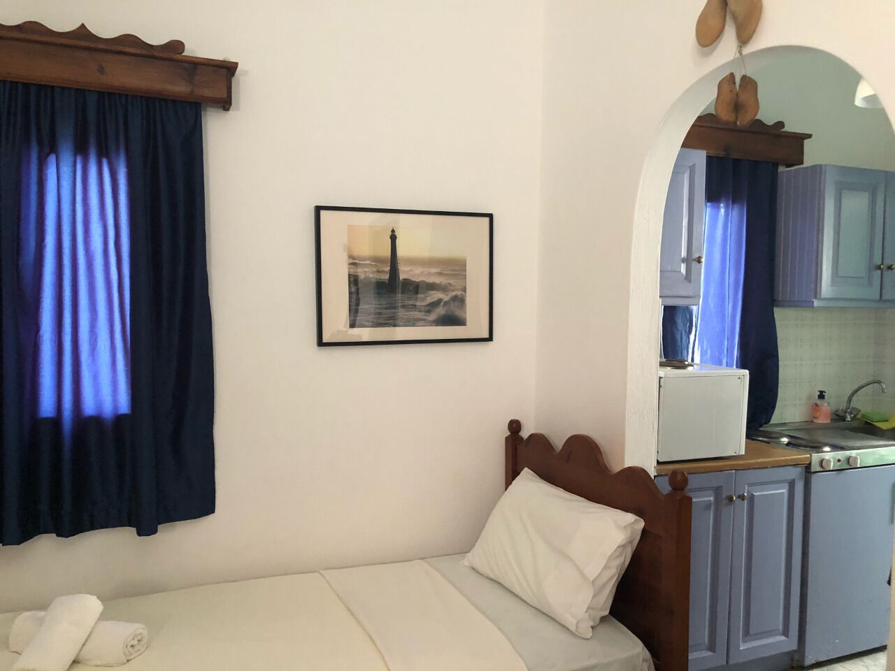 Una cama individual con cabecera de madera, un almohadón blanco y una toalla al lado. En la pared, se aprecia un cuadro enmarco con una imagen de un faro. Se observa una kitchenette con microondas, fregadero y armarios.
