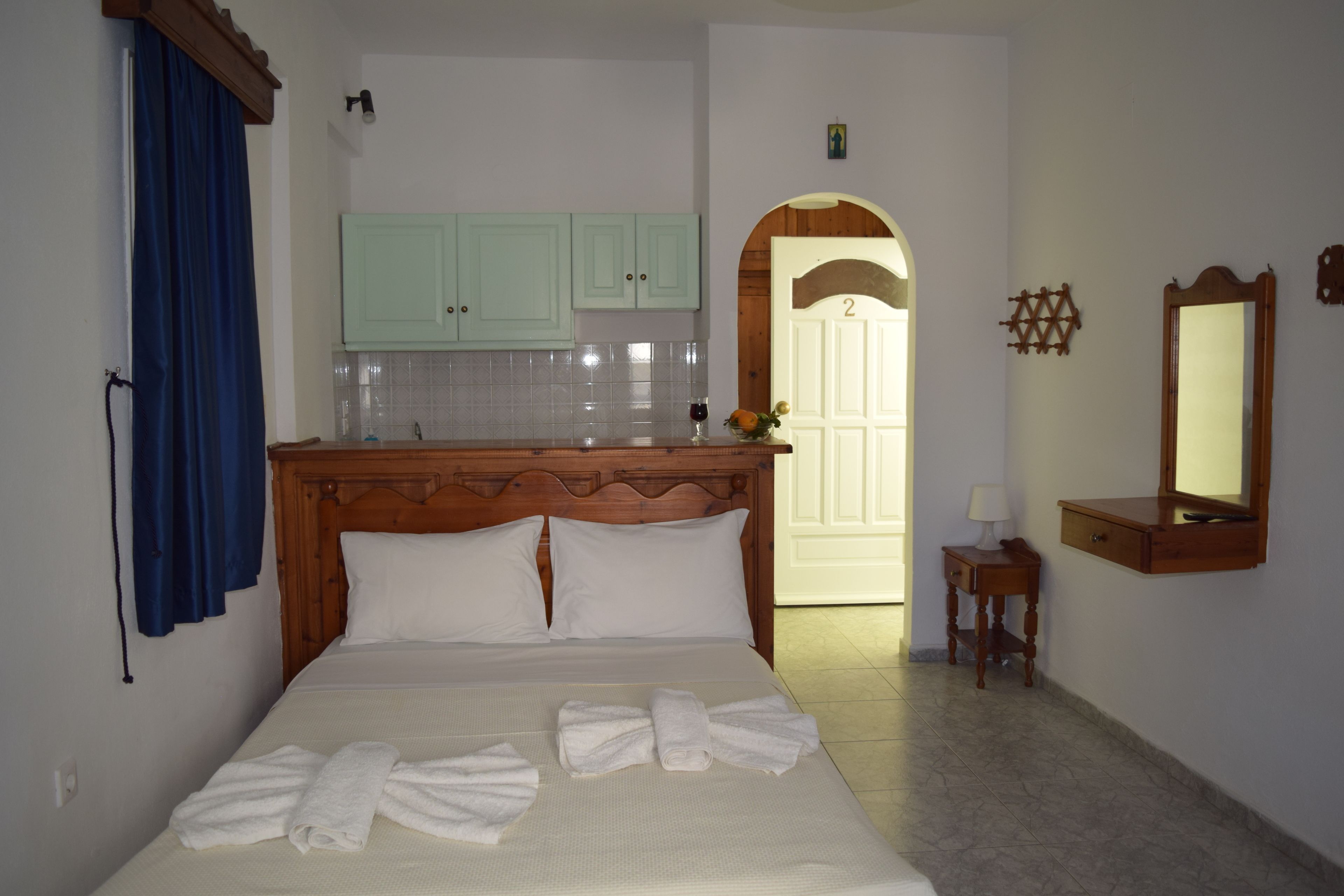 Ein Hotelzimmer mit einem Bett, einem Holzkopfende, einem kleinen Tisch mit einer Lampe, einem Spiegel und Blickrichtung zur Küche und zum Flur.