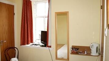 A small room with a bed, a chair, a window with curtains, a mirror, a shelf with toiletries, and a television.