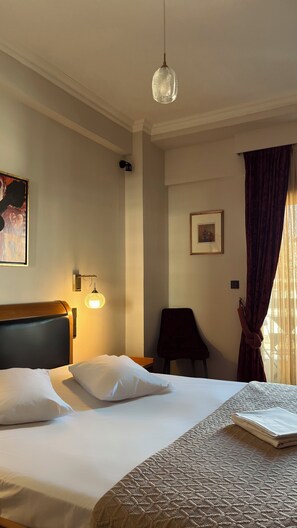 A hotel room with a bed, a headboard, a wall-mounted light fixture, a framed picture, a dark chair, and a window with curtains. - Alfa Hotel (Nikaia-Agios Ioannis Rentis)