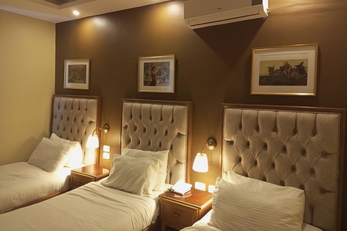 A hotel room with three beds, each with a tufted headboard, bedside lamps, and framed artwork on the walls.