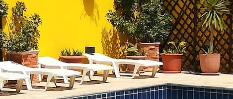 Zona de piscina con sillas de descanso blancas, plantas en macetas y una pared amarilla.