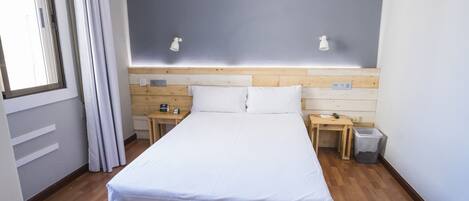 Habitación de hotel con cabecera de madera, una cama individual, dos mesitas de noche, un bote de basura y una ventana con cortinas.