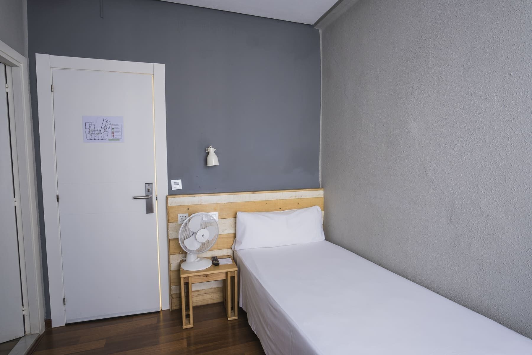 A small, single-bed room with a wooden headboard, a bedside table with a fan, a white door, and a light switch on the wall.