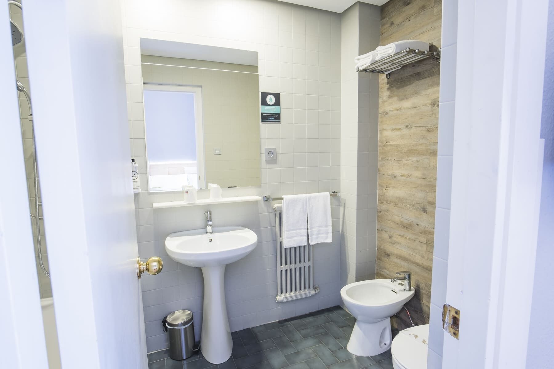 Une salle de bain avec un lavabo sur colonne, des toilettes et un bidet. On y voit un porte-serviettes avec une serviette qui y est accrochée, ainsi qu'une poubelle.