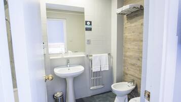 Une salle de bain avec un lavabo sur colonne, des toilettes et un bidet. On y voit un porte-serviettes avec une serviette qui y est accrochée, ainsi qu'une poubelle.
