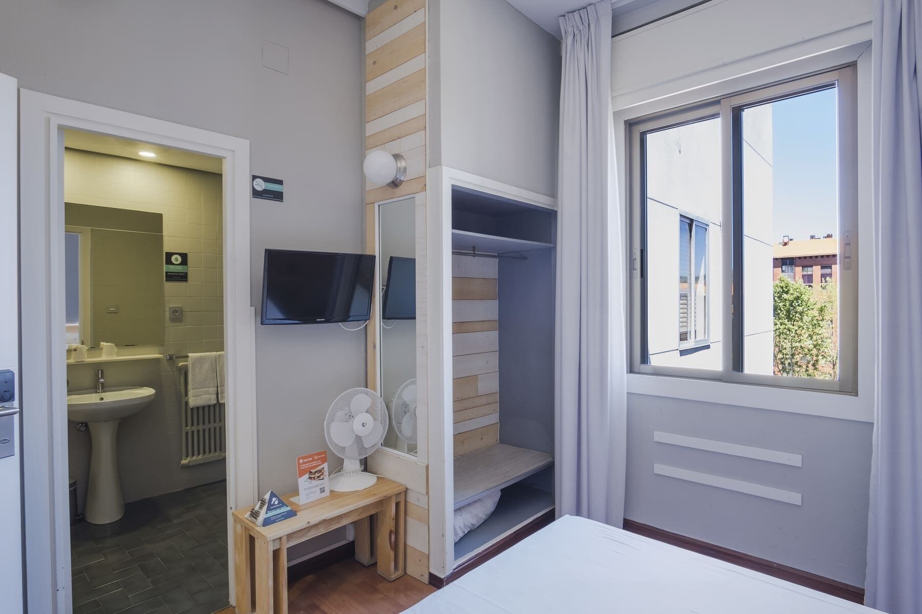 Une chambre d’hôtel de taille réduite, comprenant un lit, une petite table avec un ventilateur, un téléviseur fixé au mur et une fenêtre donnant sur les bâtiments environnants.