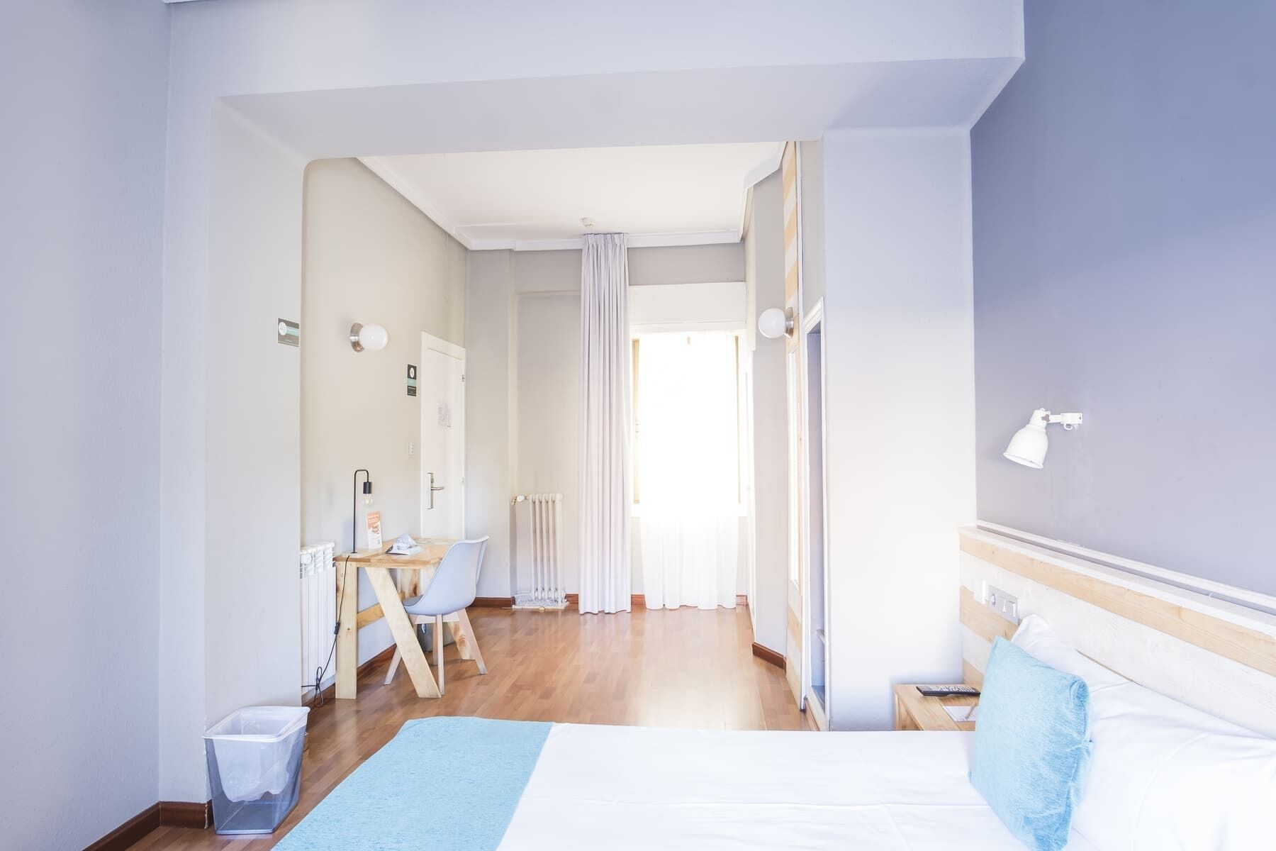 Una camera d'albergo con un letto, una scrivania con una sedia, un termosifone e una finestra con tende.