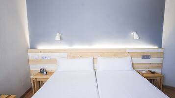 Cama doble con sábanas blancas, cabecero de madera, mesitas de noche con lámparas y un enchufe en la pared.