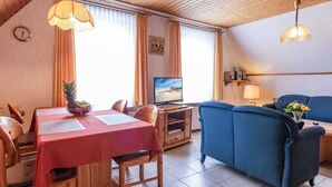 Een kamer met een eettafel, stoelen, een bank, een televisie en een klok aan de muur.
