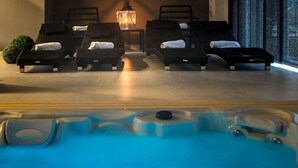 Espaço de spa com hidromassagem e quatro espreguiçadeiras, aquecedor fixado na parede e vista para árvores através de grandes janelas.