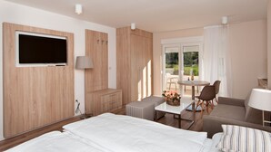 Quarto de hotel moderno com uma cama grande, uma televisão presa a um painel de madeira, uma pequena mesa de jantar com cadeiras e uma varanda com vista.