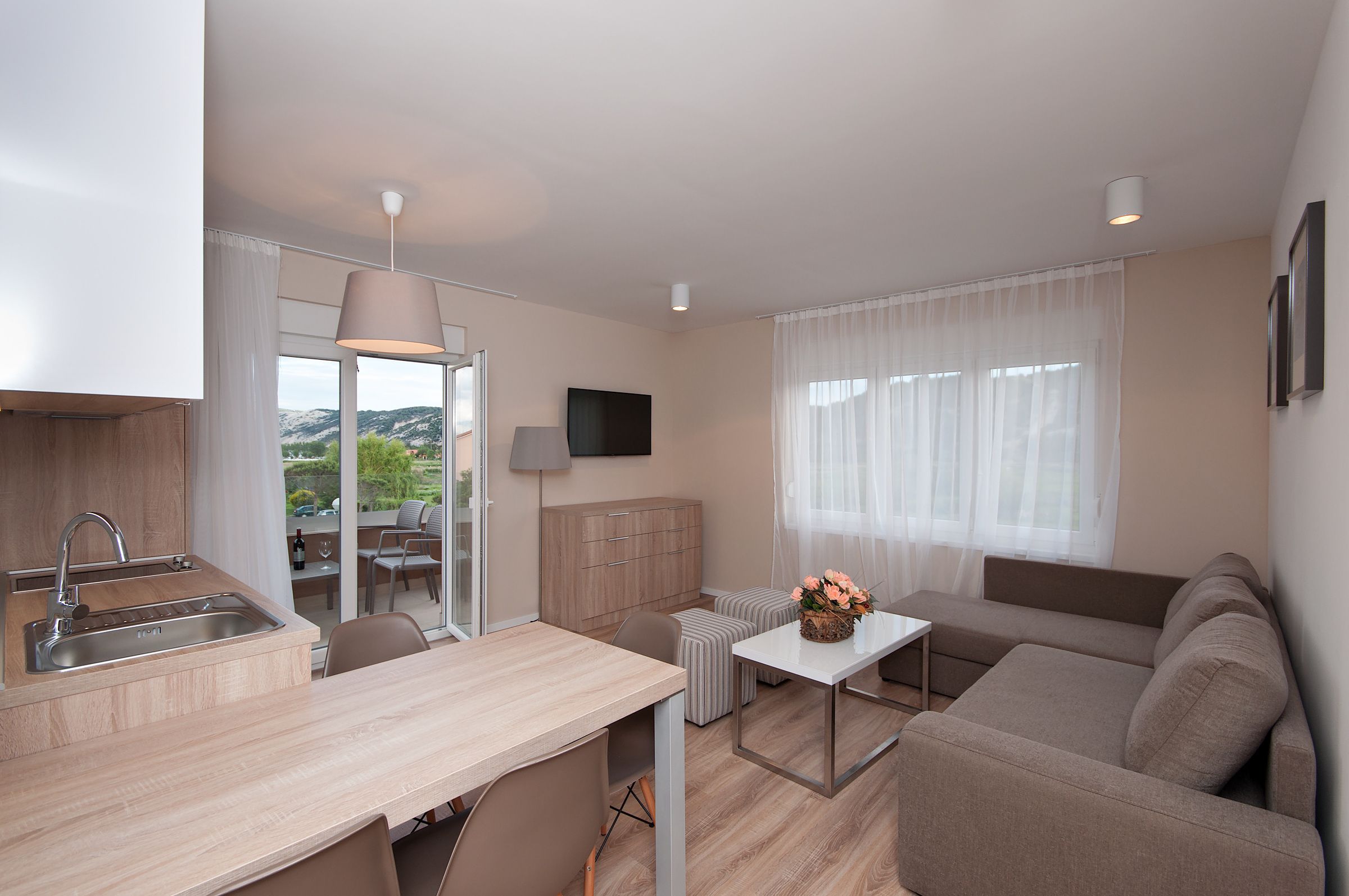 A modern kitchen with a sink, dining table, and a sofa. There is a TV mounted on the wall and a window with curtains.