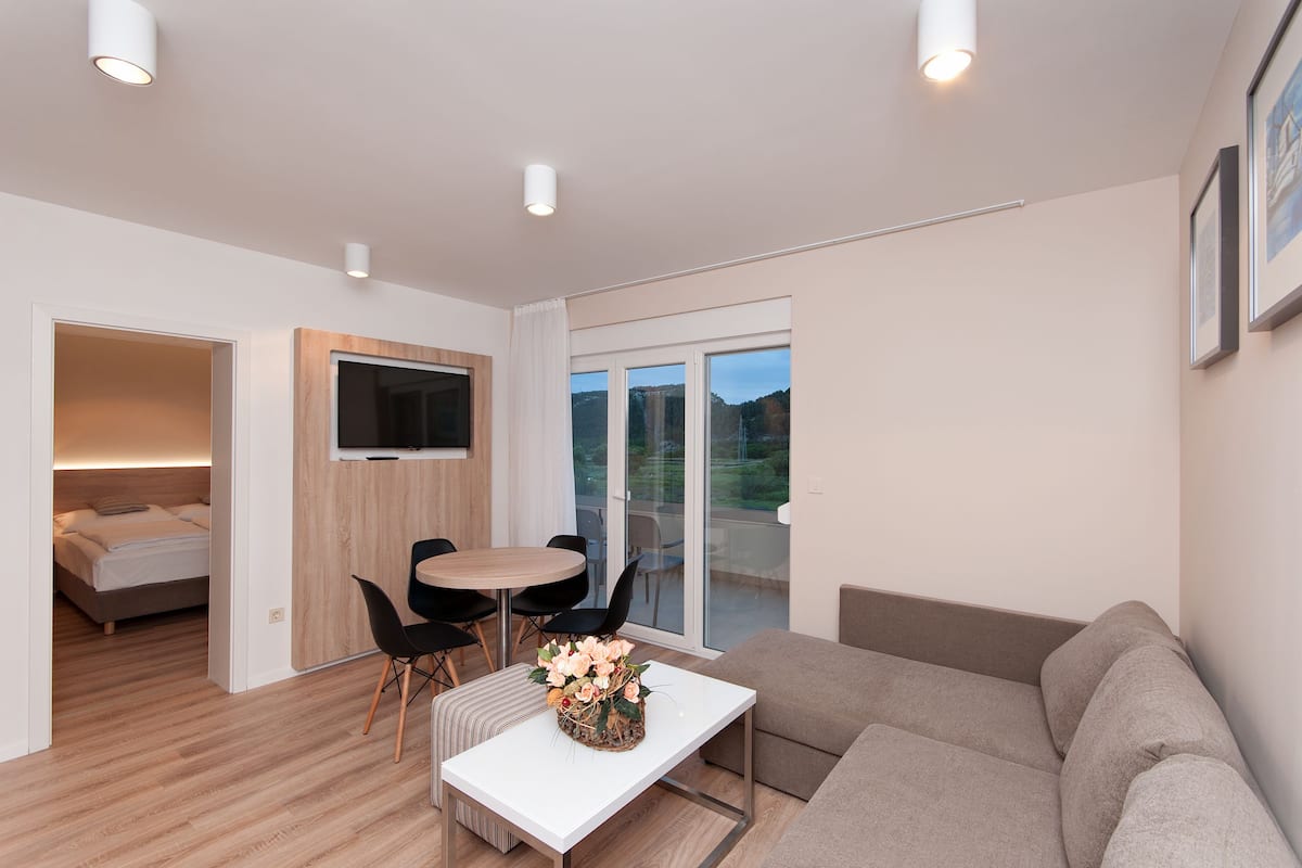 Una sala de estar moderna con un sofá, una mesa de comedor con sillas, un televisor montado en un panel de madera y un puerta corrediza de vidrio que da a un balcón.