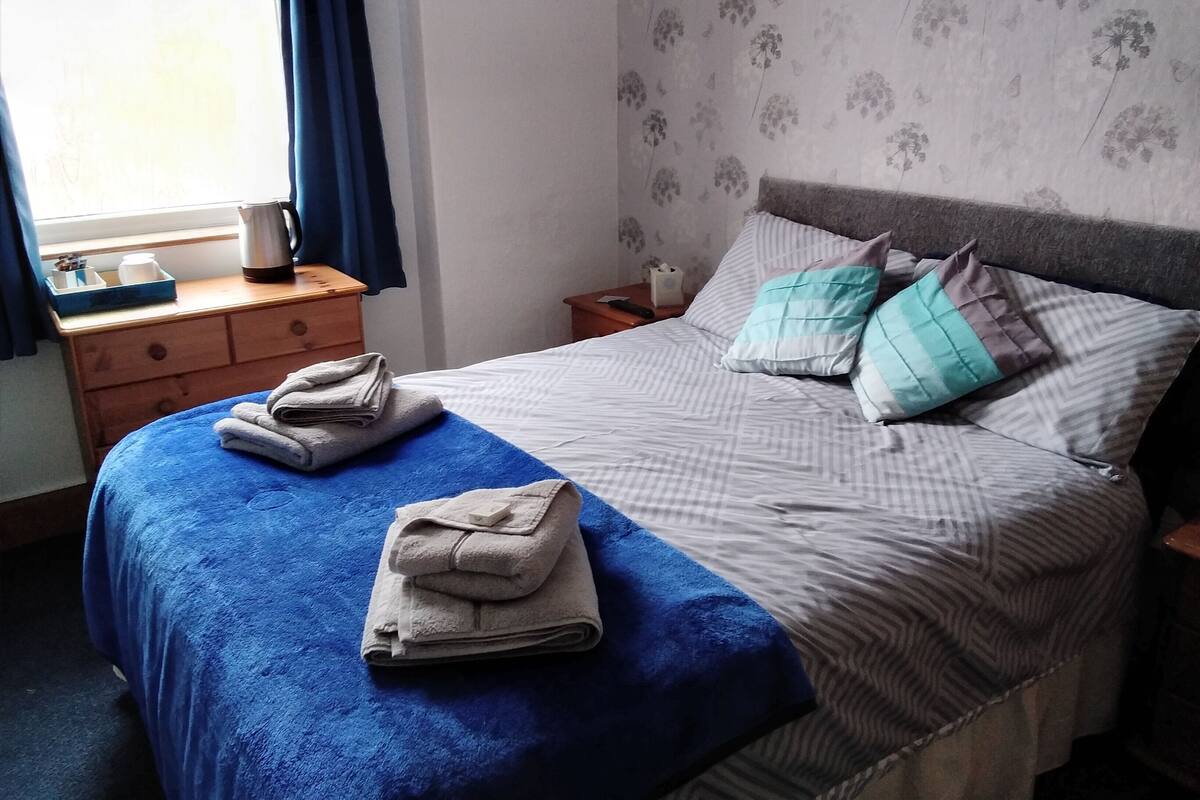 A neatly made bed with a blue blanket and folded towels, a bedside table with a kettle, and a wall with a floral wallpaper pattern.