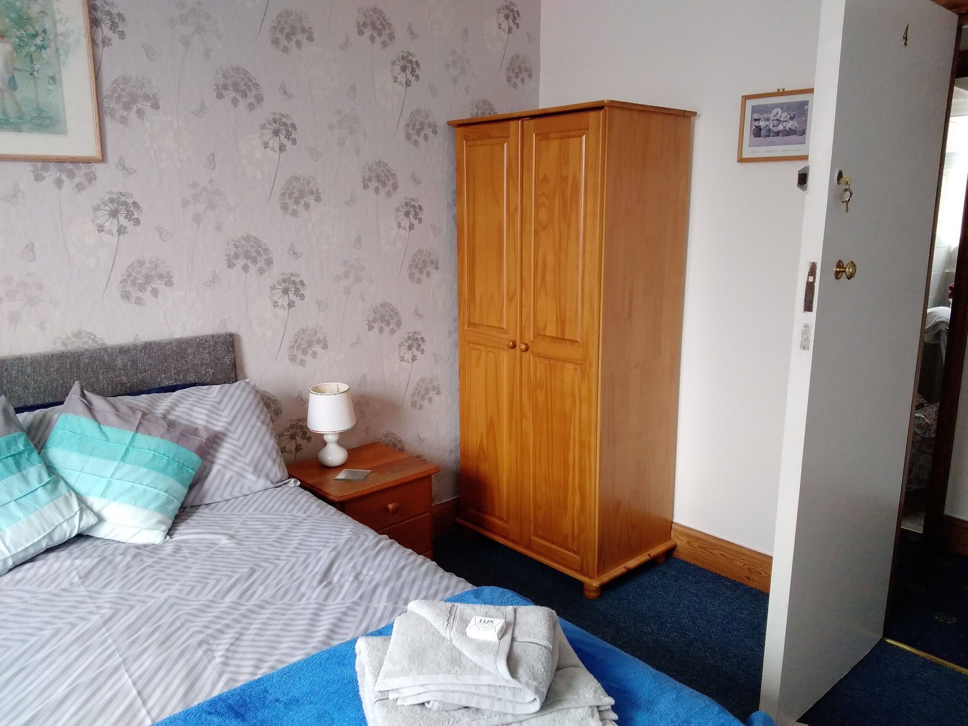 A bedroom with a bed, pillows, a nightstand, a wooden wardrobe, and a blue carpet.
