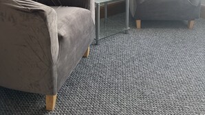 Une pièce avec un canapé gris, un fauteuil gris, une petite table en verre et un papier peint à motifs floraux.