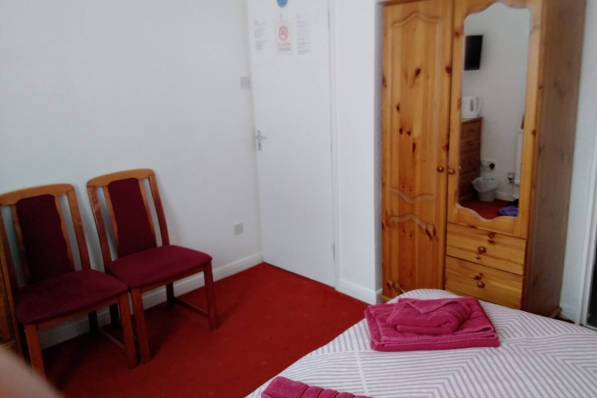 A bedroom with a bed, two chairs, a wooden wardrobe, and a door with a poster.