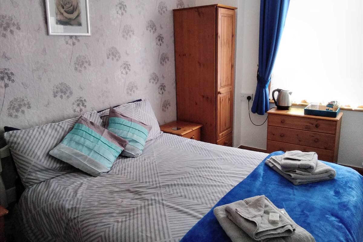 A bedroom with a bed, pillows, a wooden wardrobe, a nightstand, and a window with curtains.