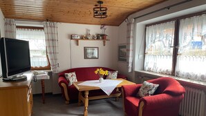 Ein gemütliches Zimmer mit Holzbalkendecke, einem roten Sofa, einem Holztisch mit Blumenvase und einem Fernseher auf einer Holzkommode.