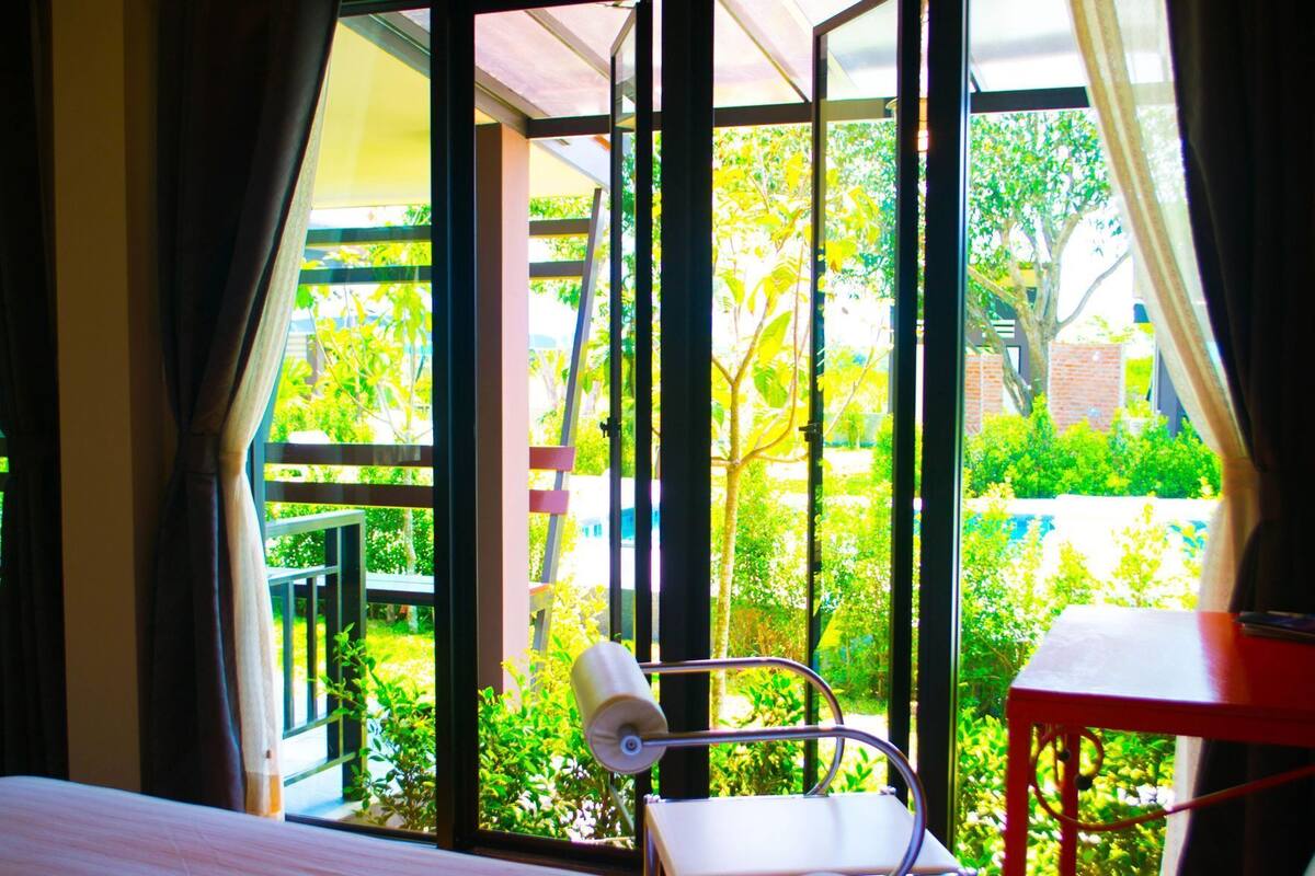 A room with a bed, a chair, a table, and a large window with a view of greenery and a building.
