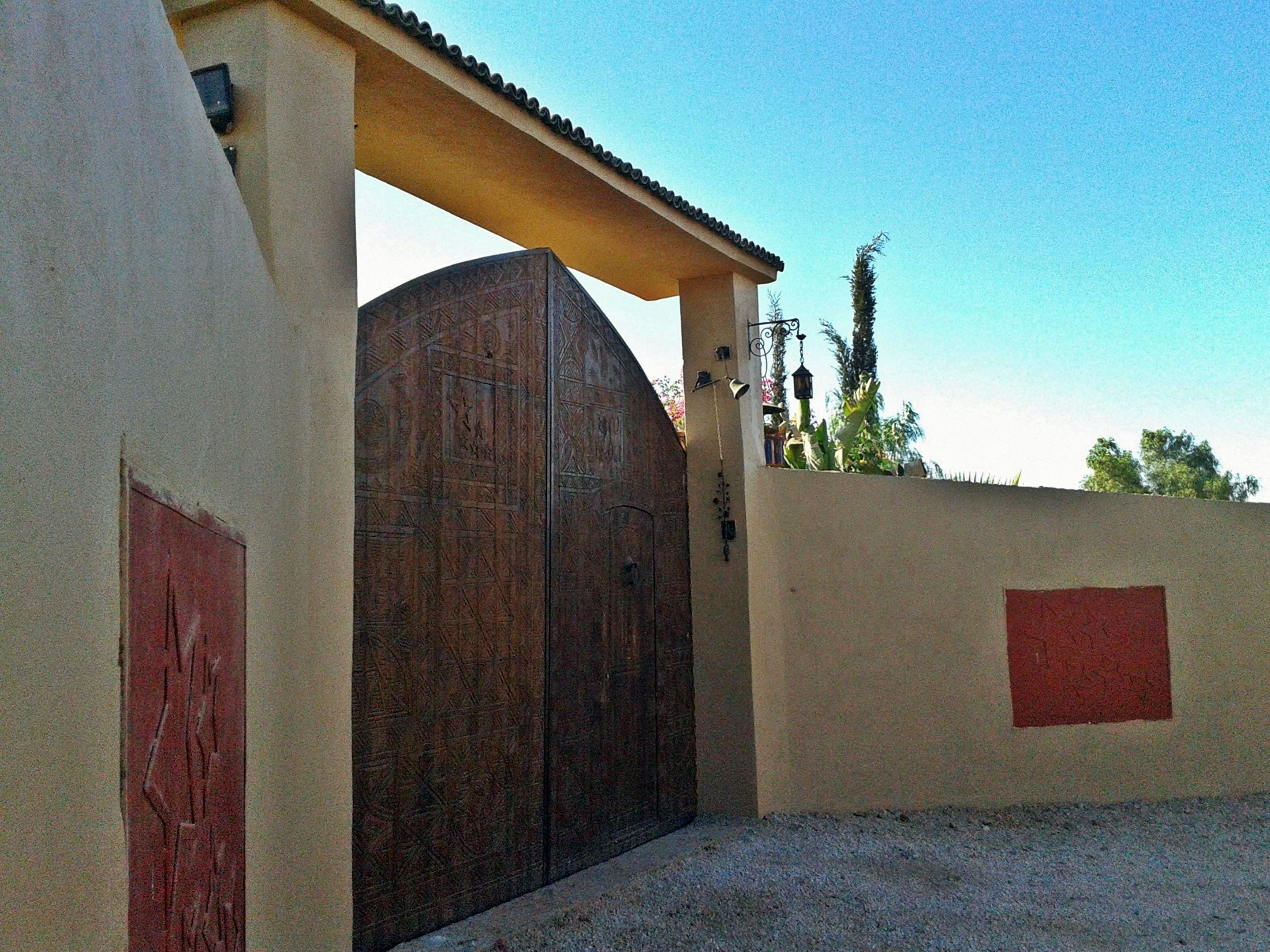 A large, ornate metal gate with intricate designs, leading to a building with a beige facade and a red rectangular feature on the wall.