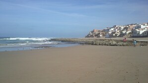 A beach with a rocky shoreline, waves crashing onto the shore, and a town with buildings in the background. - Le Jardin aux Etoiles (Sidi Boumoussa)