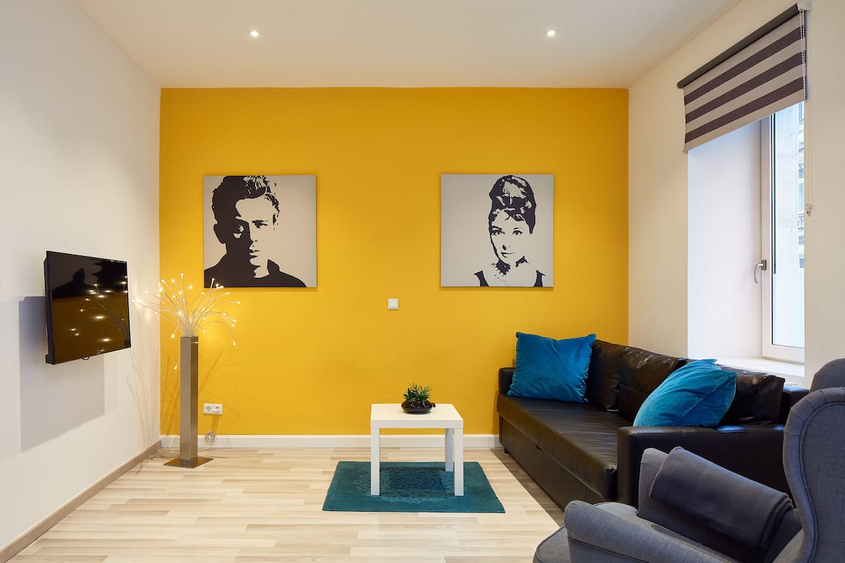 A living room with a yellow wall, two black and white portraits, a flat-screen TV, a small table with a plant, a grey sofa with blue cushions, and a window with blinds.