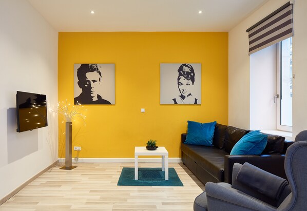 Un salón con una pared amarilla, dos retratos blanco y negro, un televisor de pantalla plana, una mesita con una planta, un sofá gris con cojines azules y una ventana con persianas.