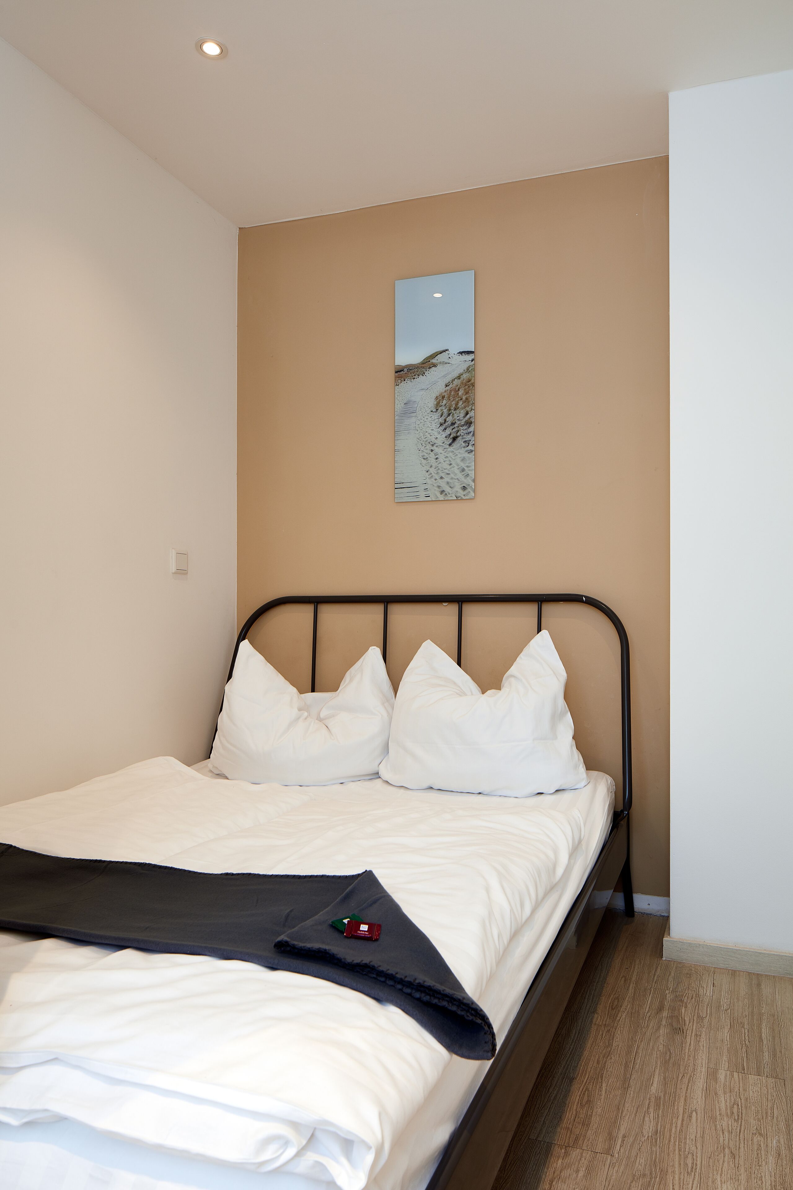A single bed with white bedding and a black blanket, a beige wall, and a framed picture above the bed.