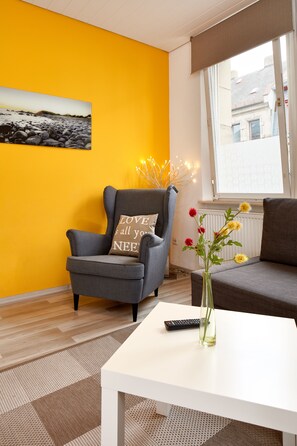 Un salón con una pared amarilla, un sillón gris con un cojín, una mesa blanca, un jarrón con flores y una ventana con vistas a edificios.
