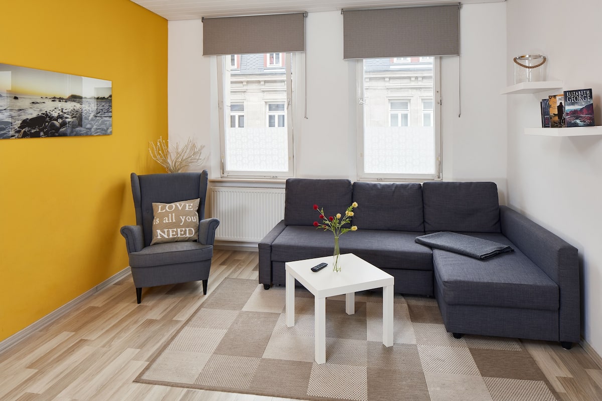A modern living room with a grey sofa, a small white table, a chair with a cushion, a painting on the wall, and a window with blinds.