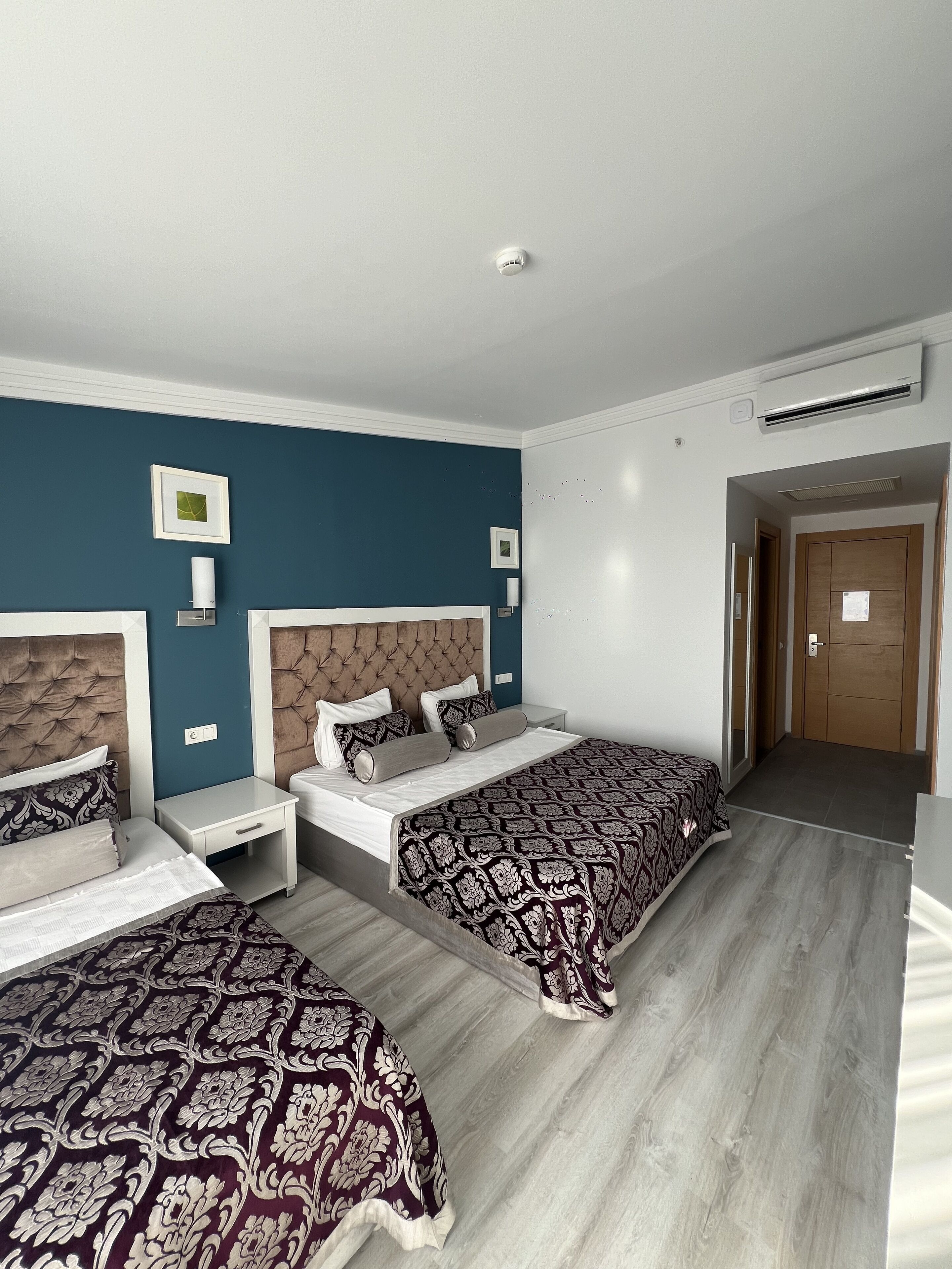 Quarto de hotel com duas camas, piso de madeira, parede azul decorativa e porta que dá para outro aposento.
