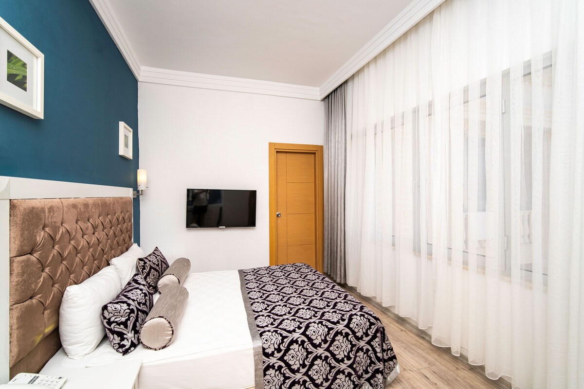 Une chambre à coucher avec une tête de lit capitonnée, un lit avec une literie à motifs, un téléviseur fixé au mur et une porte avec des rideaux.
