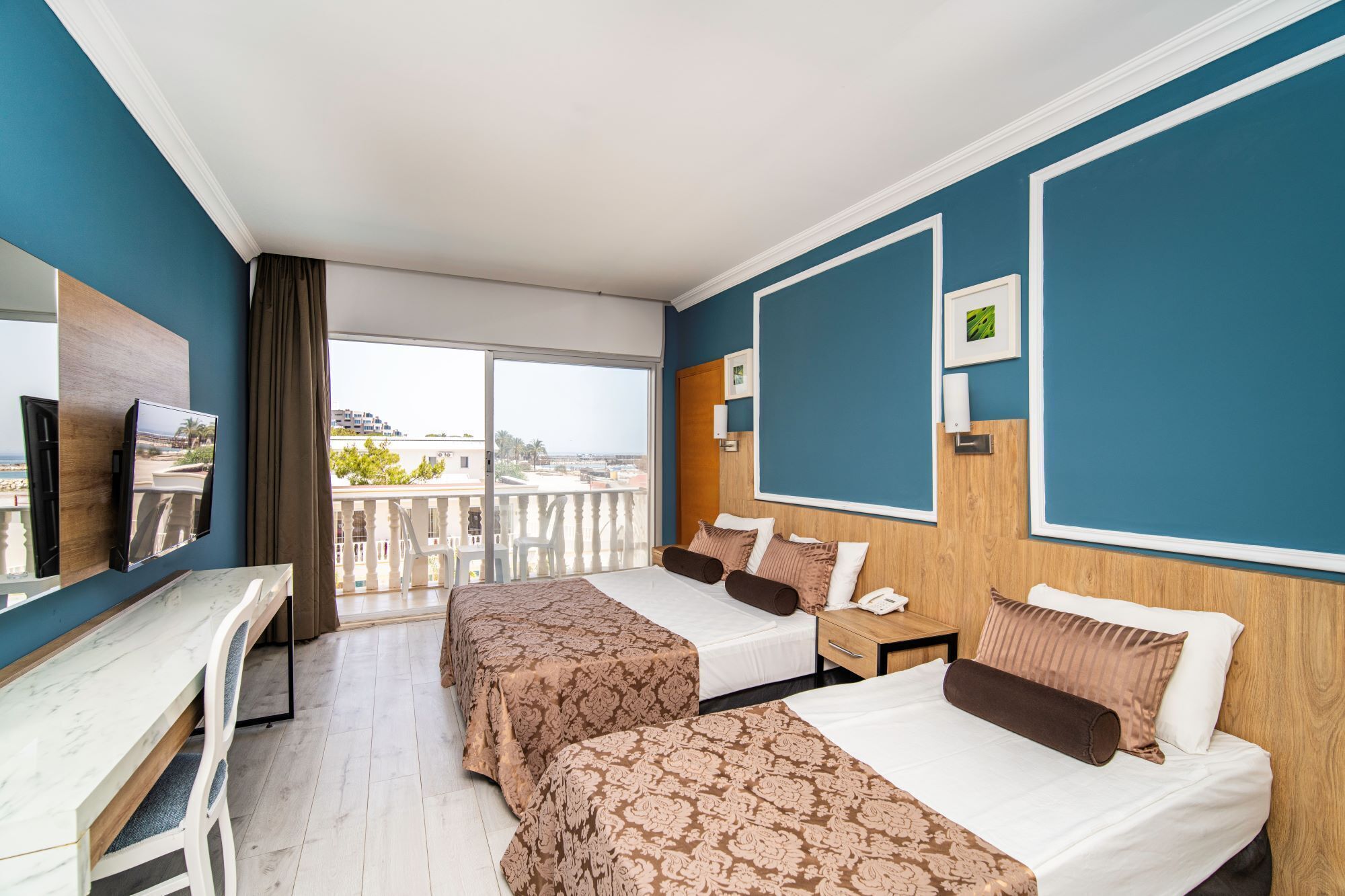 A hotel room with two beds, a desk, a TV, and a balcony with a view of the city.