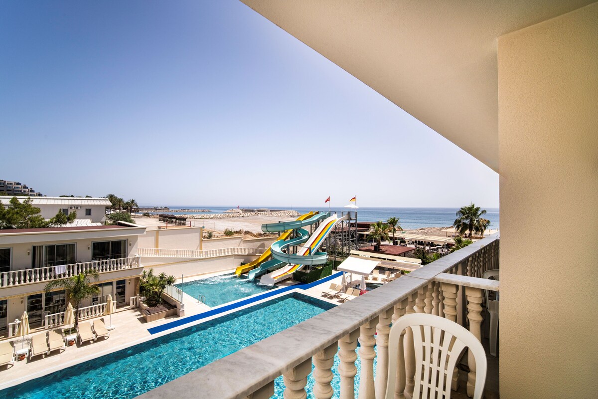 Une piscine d’un complexe hôtelier avec un toboggan aquatique, une vue sur la plage et un balcon équipé d’une chaise.