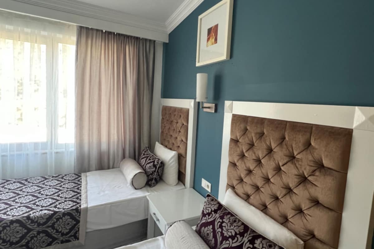 Habitación de hotel con dos camas, un ventanal con cortinas, una lámpara de pared y un cuadro enmarcado.