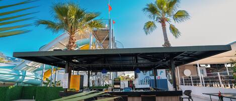 An outdoor seating area with a covered bar, palm trees, and a water slide in the background.